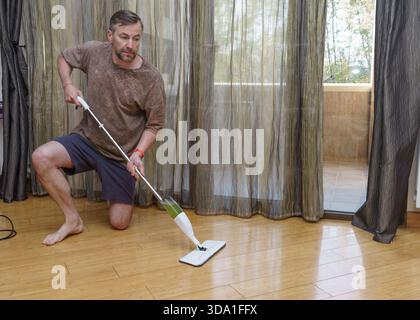 A middle-aged man with a beard, barefoot and kneeling, cleans a shiny bamboo floor with a modern spray mop in a room with sheer curtains. Stock Photo