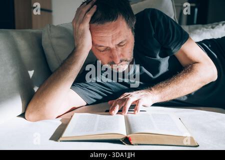 Authentic middle-aged man lying on couch, looking exhausted and deep in thought while reading an open book, hand on forehead. Perfect for overthinking Stock Photo
