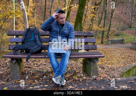 Middle-aged digital nomad working remotely on tablet while sitting on park bench in autumn forest Stock Photo