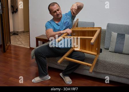 Middle-aged man assembling wooden furniture piece while sitting on floor in modern apartment living room Stock Photo
