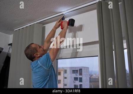 Middle-aged man using cordless drill to install curtain rod mounting bracket above window with arms raised overhead Stock Photo