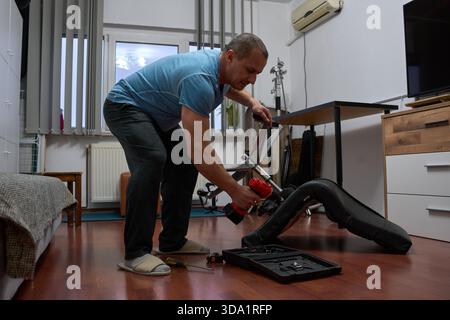 Middle-aged man repairing office chair wheels with power tool in modern apartment living room interior Stock Photo