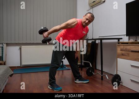 Athletic middle-aged man performing standing tricep extension with dumbbell in home gym Stock Photo