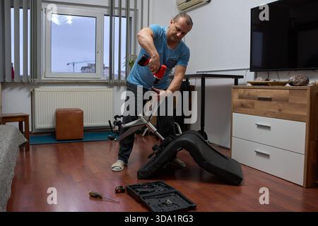 Middle-aged man repairing office chair wheels with power tool in modern apartment living room interior Stock Photo