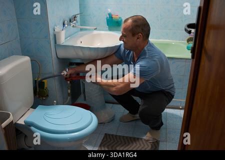 Middle-aged man using wrench to repair pipes under bathroom sink while crouching on tiled floor Stock Photo