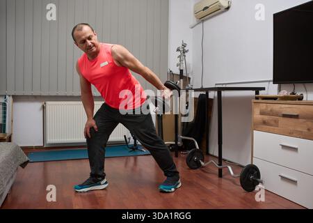 Athletic middle-aged man performing standing tricep extension with dumbbell in home gym Stock Photo