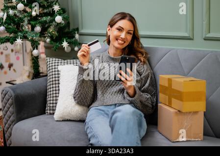 A smiling woman sits comfortably on a sofa, holding a credit card and smartphone, with delivered packages beside her and a decorated Christmas tree in Stock Photo