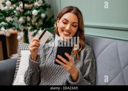 Sitting comfortably on a sofa, woman shows her credit card while using her smartphone for online shopping. Stock Photo