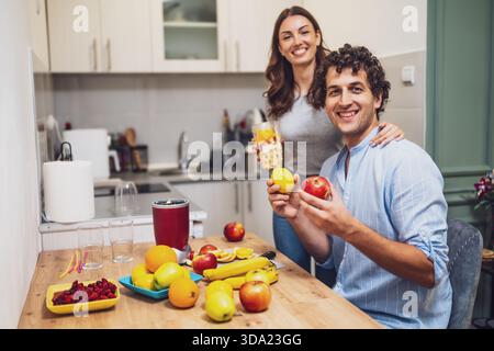Smiling and relaxed, the two share a moment together while preparing homemade fruit drinks, with a countertop full of oranges, apples, and bananas. Stock Photo