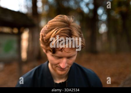 Young man with long, wavy hair looks down in a forest setting Stock Photo