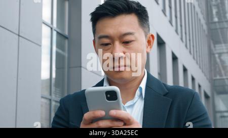 Pensive Asian Korean business man with mobile phone thinking typing message middle-aged Korean businessman in city deciding think idea browsing Stock Photo