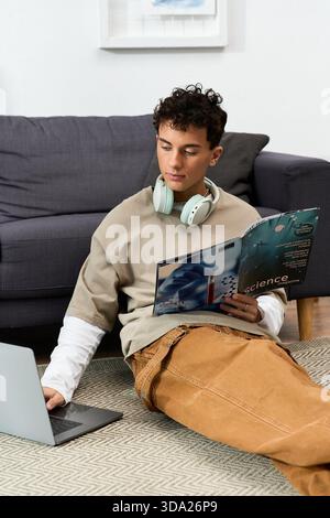 A stylish teenager relaxes on the floor at home, reading a magazine and using a laptop. Stock Photo