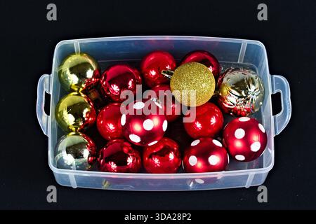 A clear plastic box of different colored Christmas ornaments sits on a table with a black background. Stock Photo