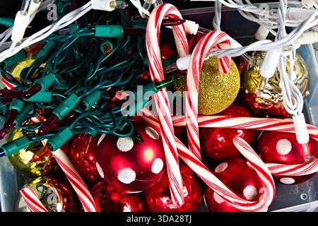 A macro shot of a plastic box with different colored Christmas ornaments, a string of lights, and some candy canes against a black background. Stock Photo