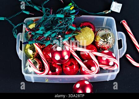 A clear plastic box with different colored Christmas ornaments, a string of lights, and some candy canes against a black background. Stock Photo