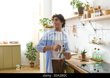A handsome young man stands in his cozy apartment, smiling as he prepares coffee. Stock Photo
