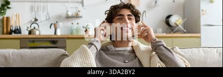 A young man with curly hair relaxes in his stylish apartment, wearing headphones and smiling, banner Stock Photo