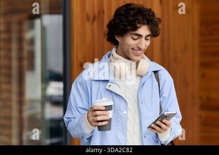 Young man with curly hair smiles outdoors, checking his phone while holding coffee. Stock Photo