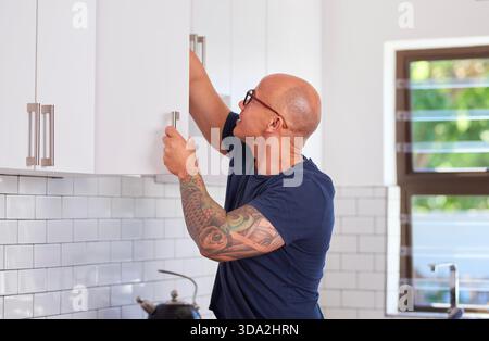 Bald Tattooed Man in Blue Shirt Opening White Kitchen Cabinets for Home Renovation Project Today Stock Photo