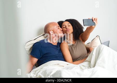Couple Kissing in Bed Taking Selfie Together in Morning Light, Cozy Love Moment Stock Photo