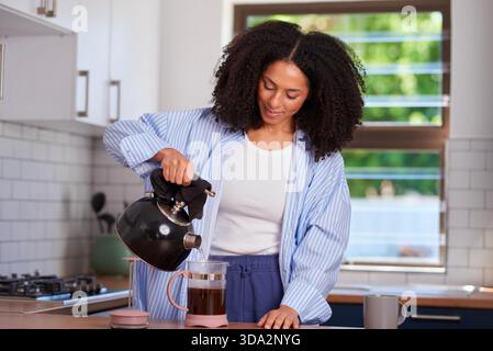 Woman Pours Hot Water From Kettle Into French Press In Cozy Morning Kitchen Stock Photo