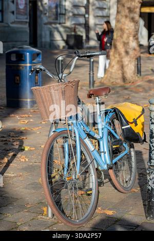 traditional vintage student style bicycle with wicker basket on the front and pannier bags locked to a bicycle rack in a busy city centre. Stock Photo