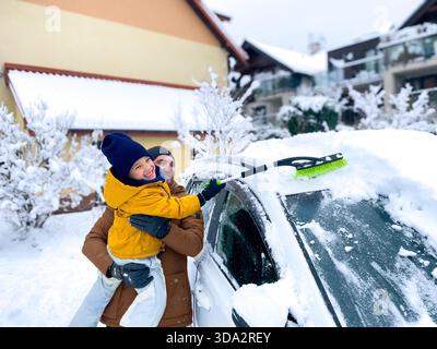 A man holds a child as they work together to remove snow from a car parked in a residential area. Snow covers the ground and surrounding houses. The s Stock Photo