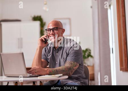 Happy Man With Glasses Works On Laptop While Talking On Phone In Modern Home Office Stock Photo