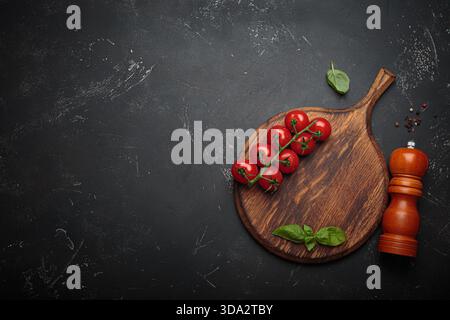 Cherry tomatoes, garlic and fresh basil leaves arranged on a rustic wooden cutting board beside a wooden pepper mill against a dark slate textured background, copy space Stock Photo