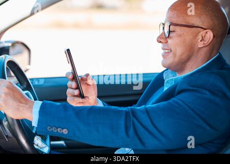 Smiling Businessman In Blue Suit Driving Car While Using Smartphone Inside Modern Vehicle Stock Photo