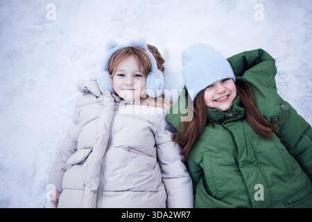 Two children are lying on snow, dressed warmly in winter jackets and earmuffs, enjoying the cold weather and creating joyful moments in a winter wonderland || Model released Stock Photo