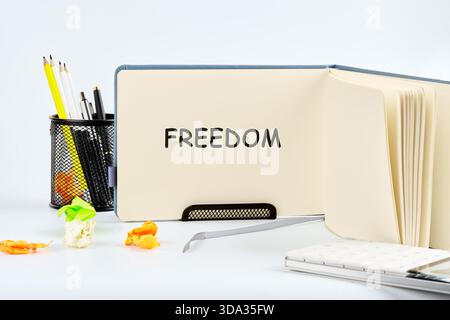 On a bright desk, a notebook lies open displaying the word freedom. Surrounding it, vibrant stationery tools and petals add a playful touch to the wor Stock Photo