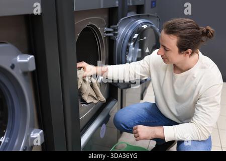 man with a bun crouching to load clothes into a washing machine at a self-service laundromat. High quality photo Stock Photo