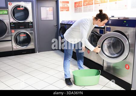 man opening the door of a washing machine at a self-service laundromat with a green laundry basket on the floor. High quality photo Stock Photo