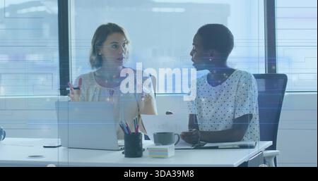 Discussing female coworkers in polka-dot and cream blouses at desk by large windows with laptop Stock Photo