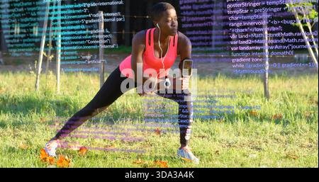Doing lateral lunge, woman in coral top stretching in park, with armband, earbuds and code overlay Stock Photo