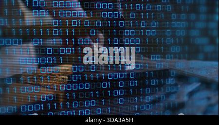 Leaning female analyst coding on laptop at dim desk, wearing dark top, glasses with binary code Stock Photo