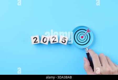 2025 Business Goals Concept. Hand holding a magnifying glass focuses on a target symbol next to wooden blocks forming the year 2025, representing futu Stock Photo