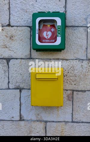 STOMORSKA, CROATIA, 10.24.2025:A green defibrillator cabinet with AED signage sits above a bright yellow mailbox on a textured stone wall, conveying p Stock Photo