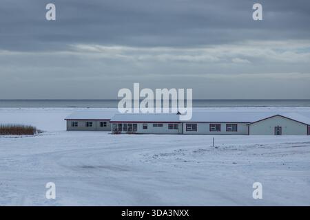 Snow covered buildings stand on a quiet coastline near Krysuvik in Iceland. Winter landscape with calm sea, overcast sky and open frozen terrain. Stock Photo