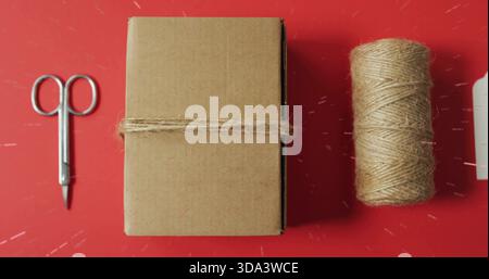 Displaying brown box wrapped with jute twine on red craft table with scissors, spool, tag Stock Photo