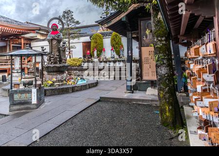 The courtyard of a Buddhist temple in Nagano, featuring a Jizo statue on a pedestal fountain, rows of small votive statues wearing red bibs, and ema wooden plaques (絵馬) hanging from a tree. Ema are small inscribed wooden tablets on which worshippers write their wishes and prayers. Nagano, Nagano Prefecture, Japan Stock Photo