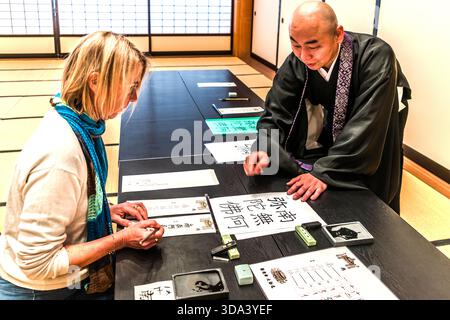 A Western woman is learning the Buddhist art of Sakky�� calligraphy (д�U, sutra copying) under the guidance of a monk in a tatami room at a sub-temple of Zenkoji in Nagano; in front of her lie brushes, an inkstone, practice sheets and a large calligraphic scroll. Nagano, Nagano Prefecture, Japan Stock Photo