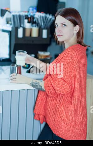 Female barista in vibrant orange sweater pours milk into pitcher at modern cafe counter with various tools and items in view. Stock Photo