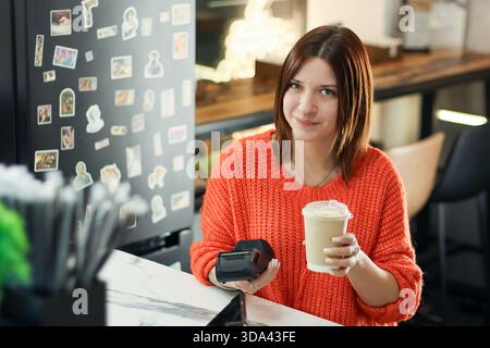 A smiling young woman in an orange sweater sits at a cafe counter, holding a takeaway coffee cup in one hand and a payment terminal in the other. Stock Photo