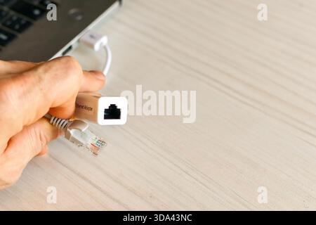 Hand grasping a white USB to Ethernet adapter linked to a silver laptop on a wooden surface with an Ethernet cable connected, showcasing portable netw Stock Photo