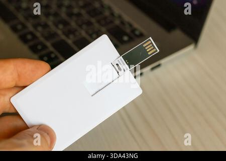 Hand positioning a white card USB flash drive with extended plug towards a laptop port for connection. Stock Photo