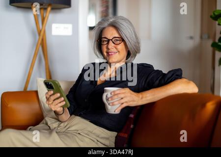Smiling mature woman enjoying a relaxing moment on the sofa, using her smartphone and drinking a cup of coffee Stock Photo