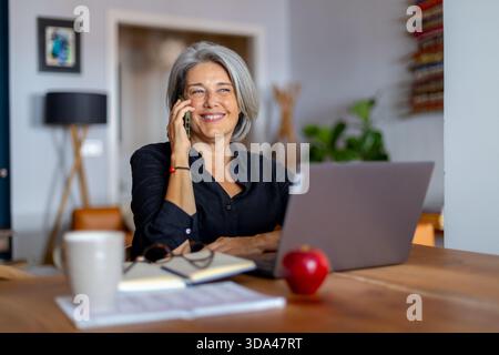 Grey haired mature businesswoman talking on smartphone and using laptop while working remotely at home Stock Photo