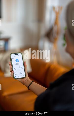 Woman holding smartphone and managing smart home devices using mobile application, controlling lights, temperature, and security systems remotely Stock Photo